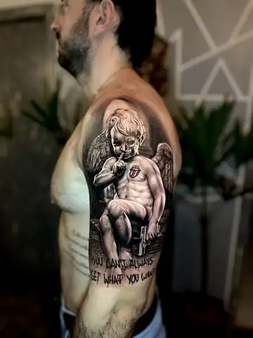 Anjo querubim com meia caveira e frase, tatuagem em realismo preto e cinza no ombro