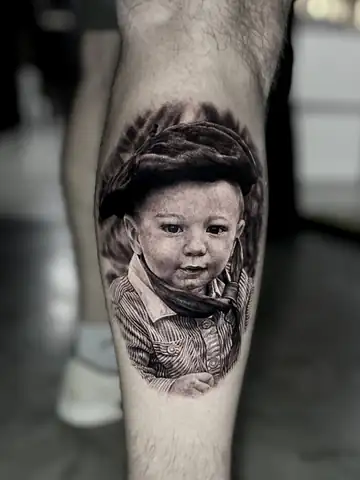 Retrato de bebê com chapéu e lenço gaúcho, tatuagem premiada em realismo preto e cinza na panturrilha