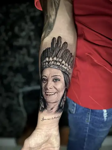 Retrato de mulher sorrindo com cocar de penas e assinatura, tatuagem de homenagem no antebraço